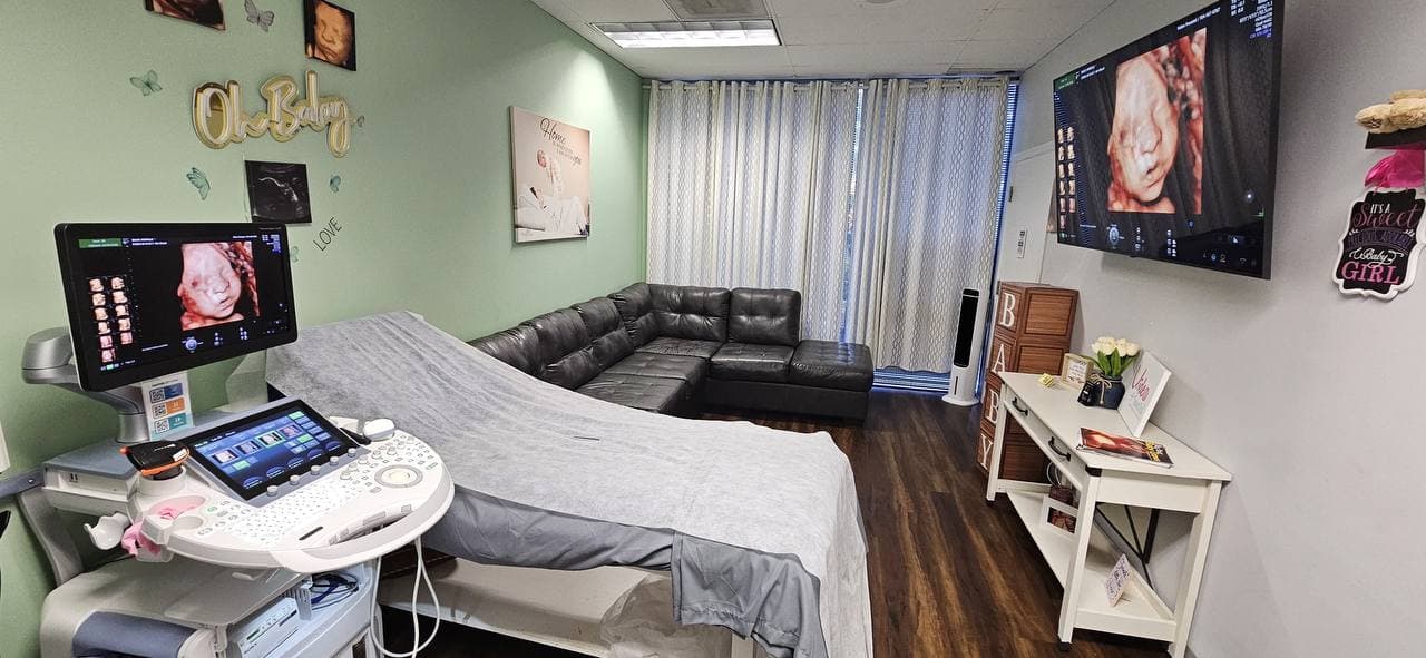 Video Prenatal ultrasound room in Charlotte with the GE Voluson ultrasound machine, scanning bed, large wall screen with a 3D baby image and a sofa for family guests