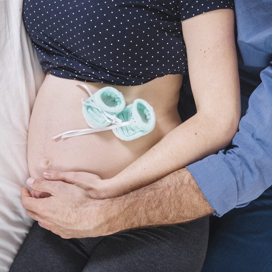 Expecting couple resting on a bed with tiny knitted baby booties placed on the pregnant belly