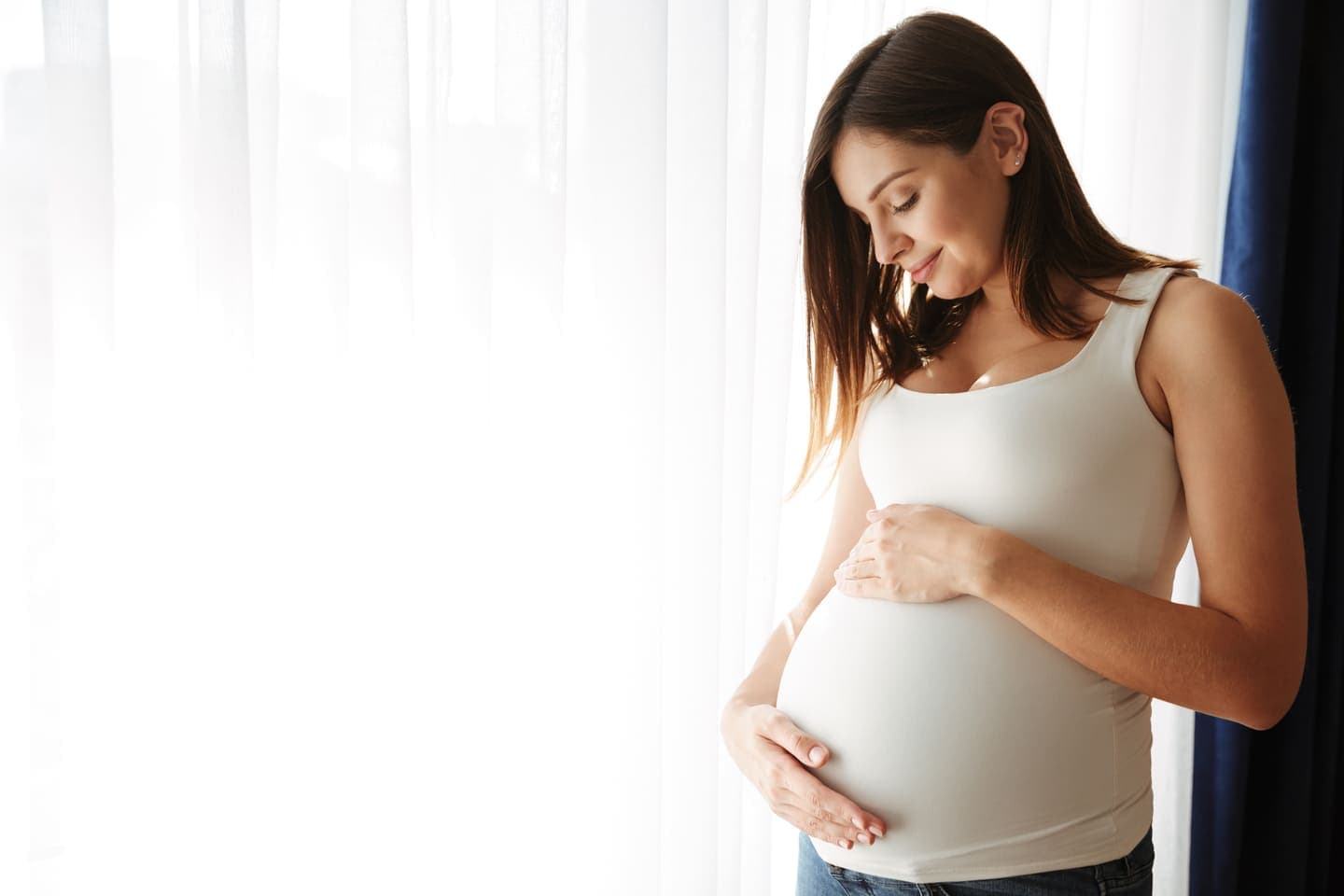 Smiling pregnant woman in a white tank top gently holding her belly by a bright window