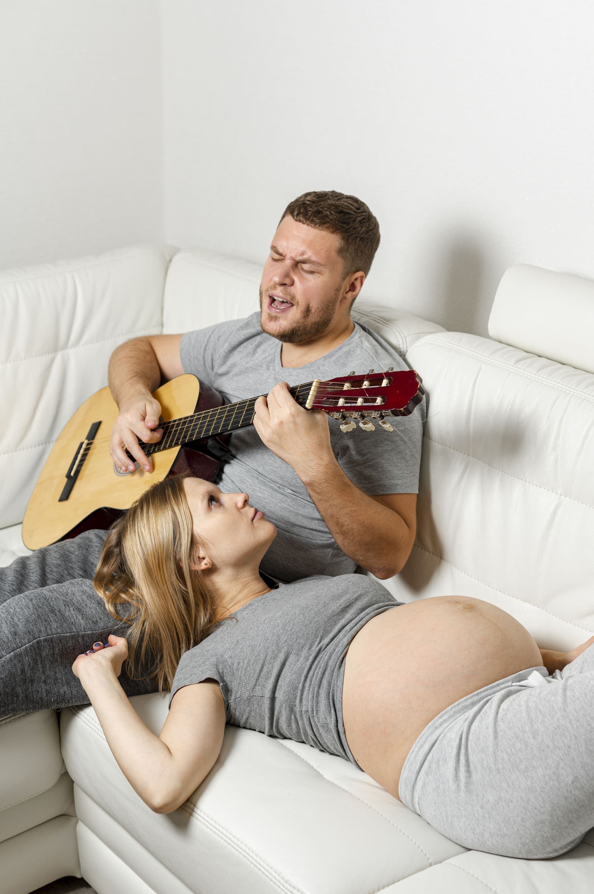 Pregnant woman resting on a white sofa while her partner plays the guitar and sings to the baby