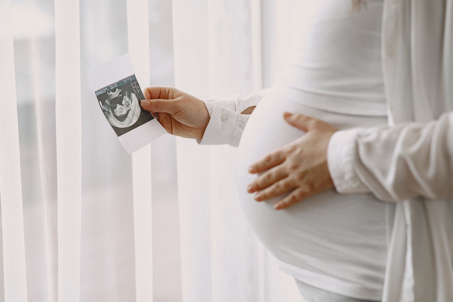 Pregnant woman in white holding a printed ultrasound photo next to her belly by a window