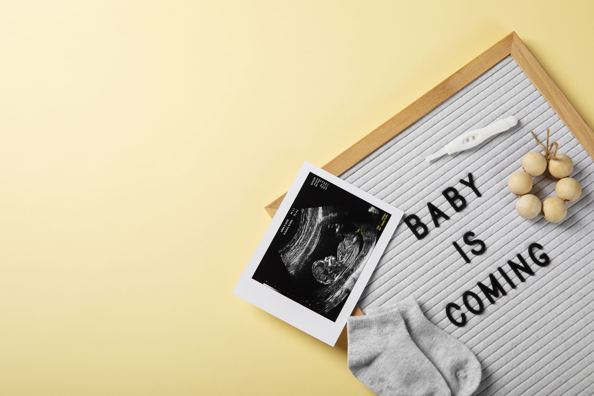 Letter board reading baby is coming next to an ultrasound print, a pregnancy test, baby socks and a wooden teether