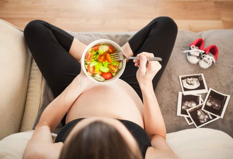 Pregnant woman seen from above eating a bowl of fruit next to printed ultrasound photos and tiny baby shoes