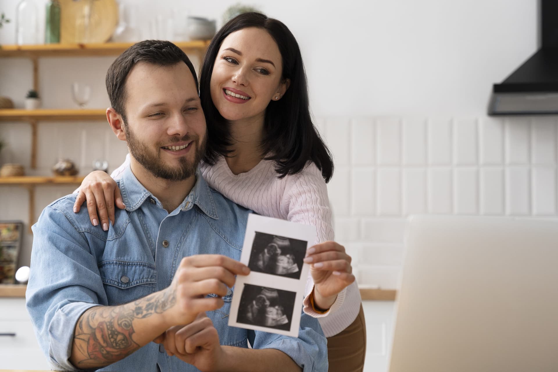 Smiling couple in their kitchen holding printed ultrasound pictures of their baby